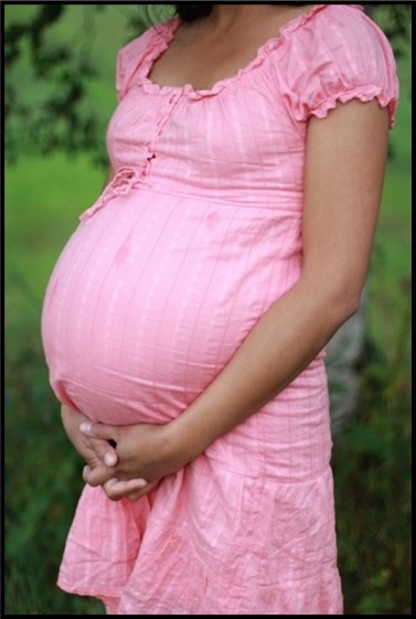 Image: A pregnant woman wearing a pink dress is shown from her neck to her knees.  She has her hands folded together beneath her belly. 