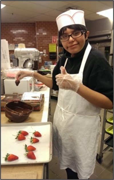 {Image: A young man is wearing a white apron and white baker’s hat.  He is standing in front of a tray of strawberries and is holding a chocolate covered strawberry in his hand over a bowl of chocolate.  He is looking at the camera smiling and he is giving the “thumbs up” sign.} 