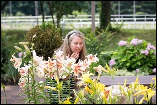 {Image: A woman is standing in front of a garden of flowers.  She has her right hand by her face and she is leaning over to smell the flowers.  There are yellow flowers in the foreground, and lavender flowers, grass, and bushes in the background.} 