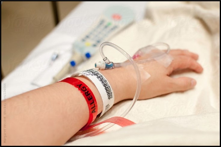 {Image: A arm and hand with IV tubes in it is lying at a person’s side.  A red bracelet that says allergy and a white bracelet with black markings are around the arm.  There is a hospital bed remote control laying next to the arm.} 