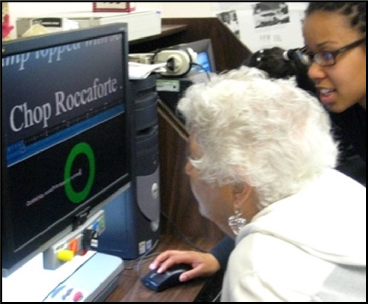 Image: A woman with white hair is sitting in front of a desktop computer screen.  She is leaning in close to the screen and the words on the screen are very large.  Another woman is sitting beside her looking at the screen and she has her hand on the computer mouse.