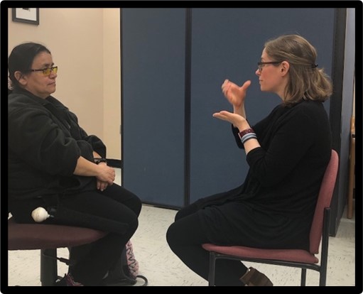 Two woman seated facing one another approximately 10 inches apart. One woman is signing. 