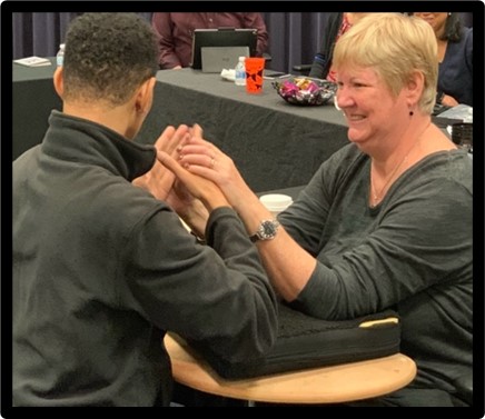 A man and woman are facing each other. The man is signing and the woman has both hands over the signer's hands