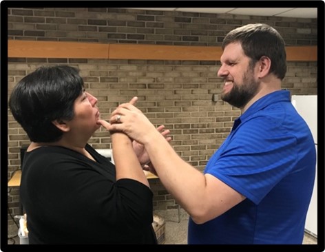 A man and woman are facing each other. The woman is signing. The man has his left hand over the signers hands and he is smiling.